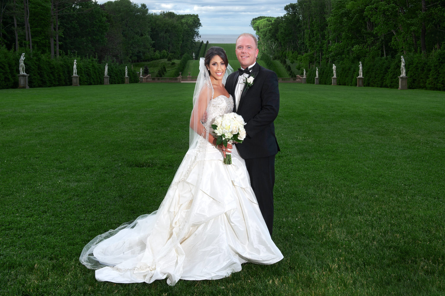 A bride and groom pose on a vast, manicured lawn flanked by statues, with the expansive greenery and distant trees creating a grand backdrop. ​