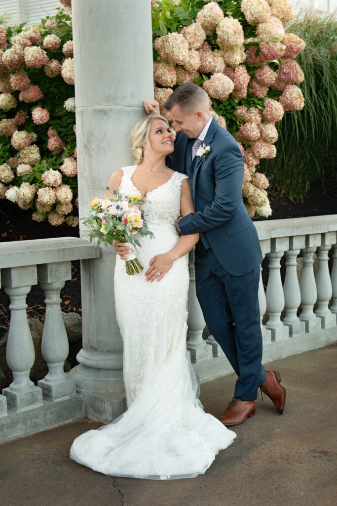 A bride and groom share a romantic moment by a column adorned with blooming hydrangeas, with the bride holding a bouquet and looking up at the groom.