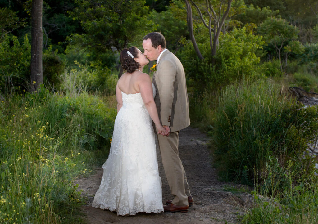 A couple shares a kiss while holding hands on a forested path, surrounded by lush greenery, in an intimate outdoor setting.