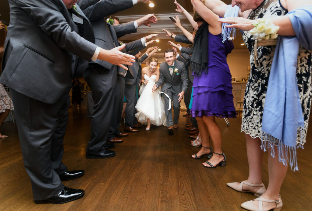 Newlyweds are joyfully running under an archway formed by their wedding party's arms, capturing a lively and celebratory moment indoors.