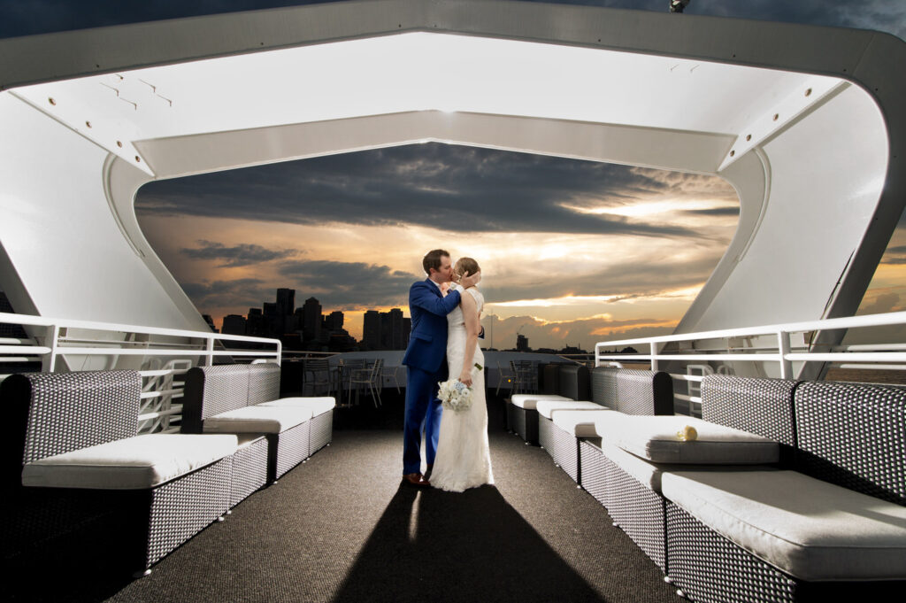 A couple kisses on the deck of a yacht at sunset, with the Boston skyline silhouetted against a dramatic sky in the background.