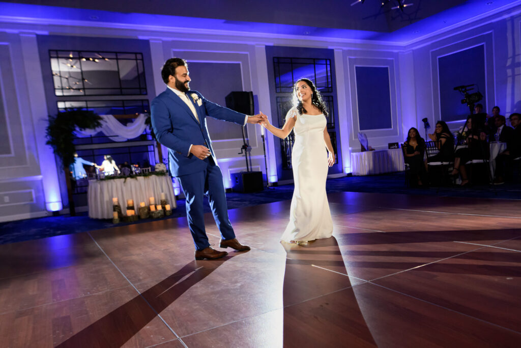 A bride and groom share their first dance on a dimly lit dance floor, surrounded by guests, with elegant decor and lighting in the background.