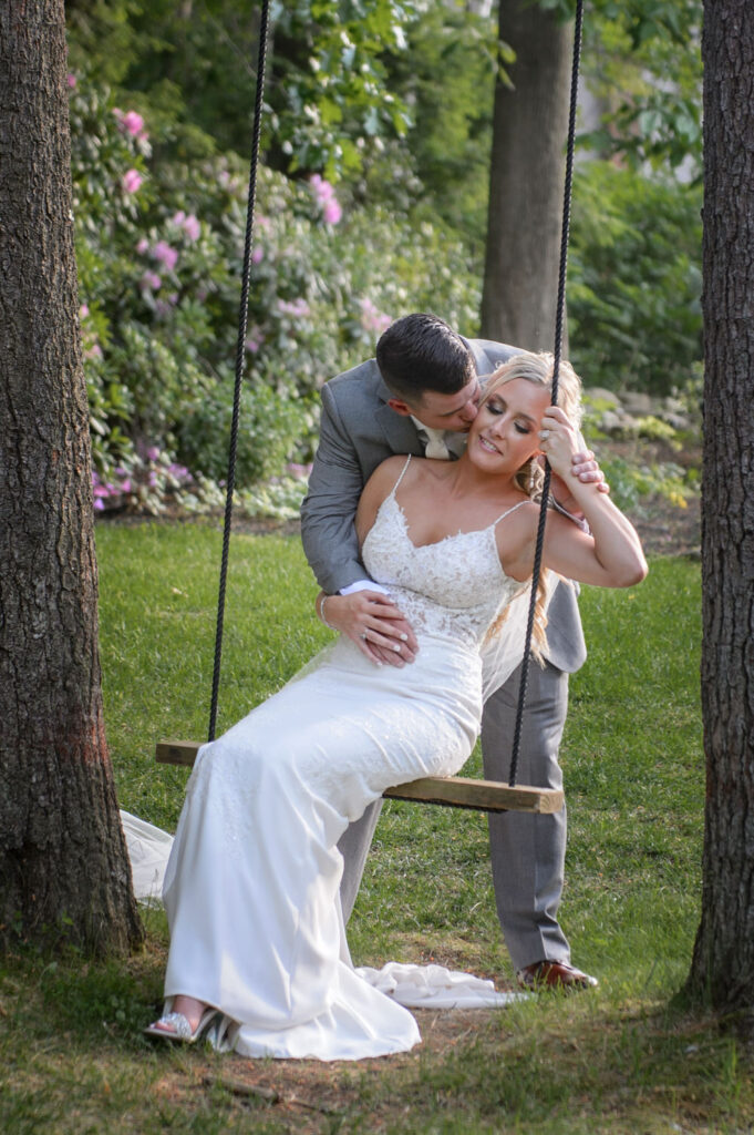 A groom kisses the bride on the cheek as she sits on a swing, with the verdant garden in the background, capturing a playful and tender moment.