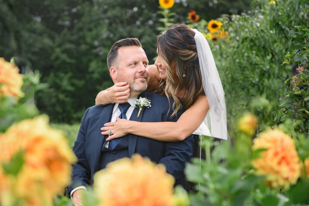 A bride embraces her groom from behind, smiling warmly at him, with vibrant sunflowers and greenery creating a picturesque outdoor setting.