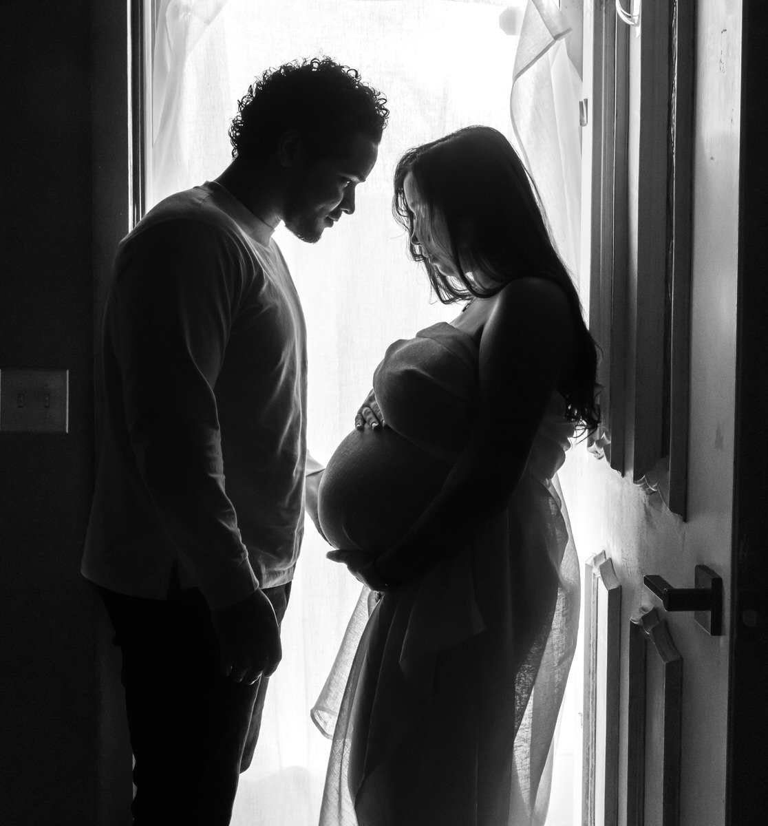 A silhouette of a couple standing face-to-face near a window, with the pregnant woman holding her belly and the man gently touching her showing what to wear for maternity photos