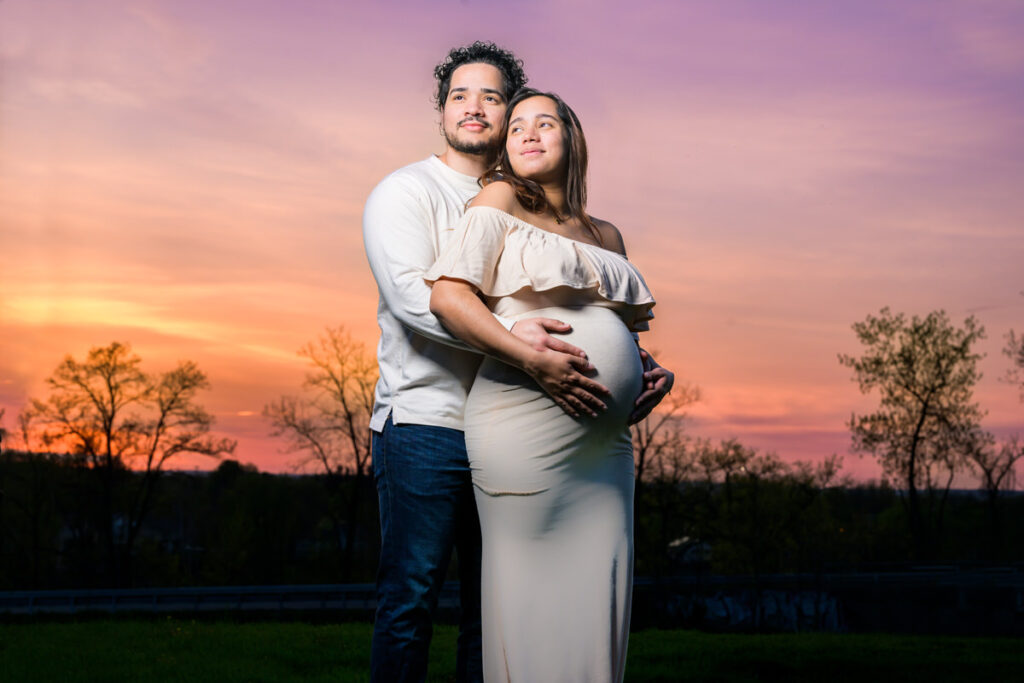 A couple standing together during a vibrant sunset, with the man embracing the pregnant woman from behind and both looking into the distance.