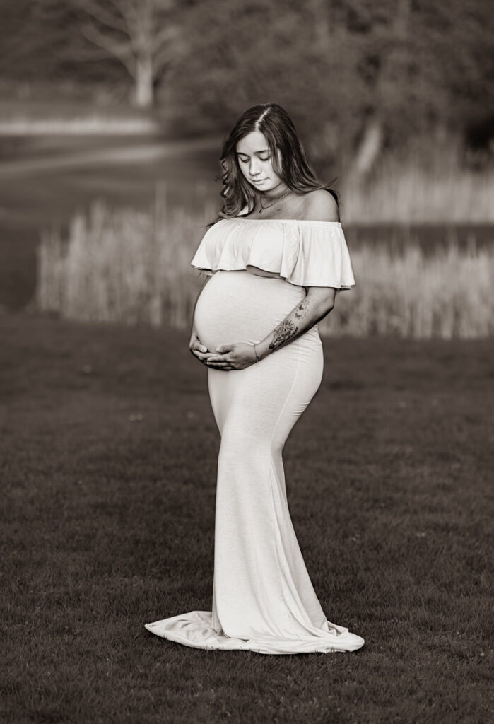 A pregnant woman in an off-shoulder dress standing alone in a grassy outdoor setting, looking down and cradling her belly.