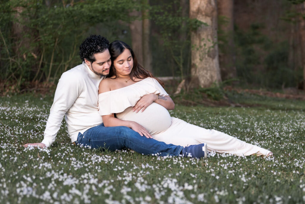 A couple sitting on the ground in a field of small white flowers, with the man resting his head on the pregnant woman's shoulder. ​​