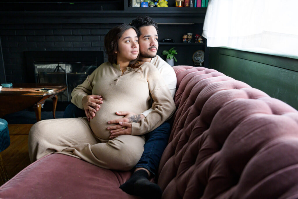 A couple sitting on a couch, with the man embracing the pregnant woman from behind, both looking out a window.