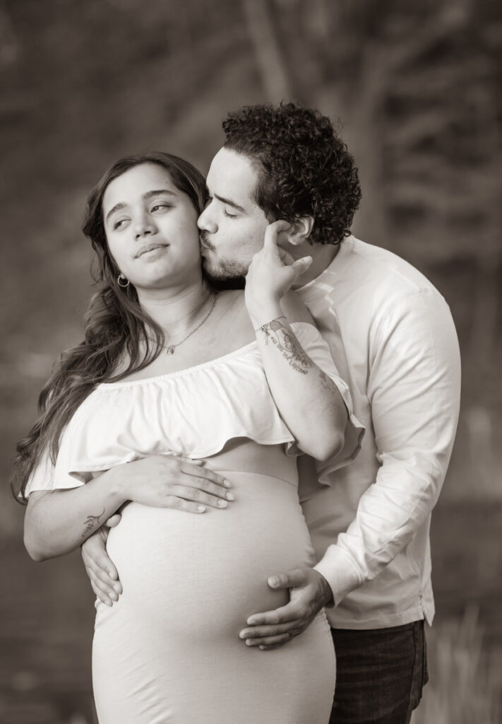 A man kissing a pregnant woman's cheek while holding her belly from behind, both standing outdoors.