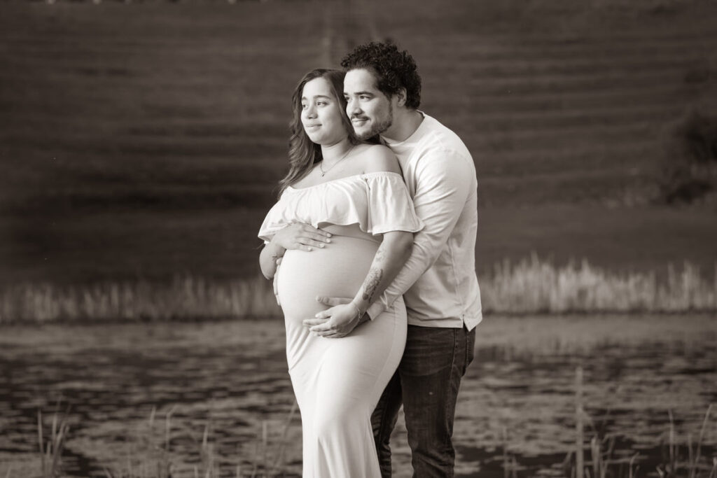 A couple standing by a lake, with the man embracing the pregnant woman from behind, both looking into the distance.