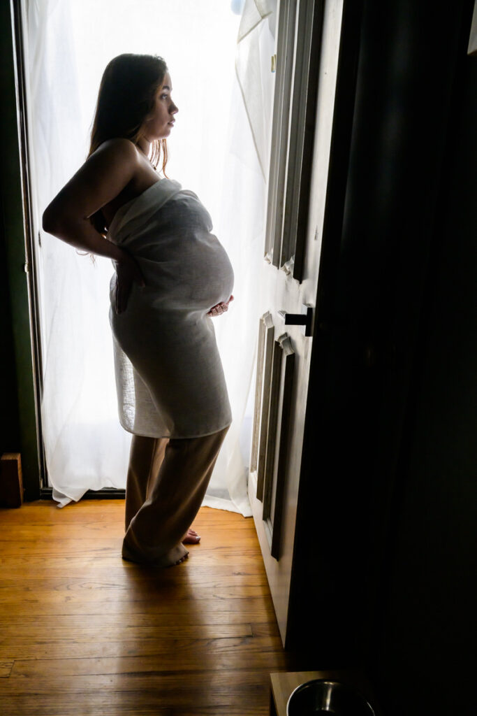 A pregnant woman standing in profile near a window, wrapped in a light cloth, with her hand on her lower back.