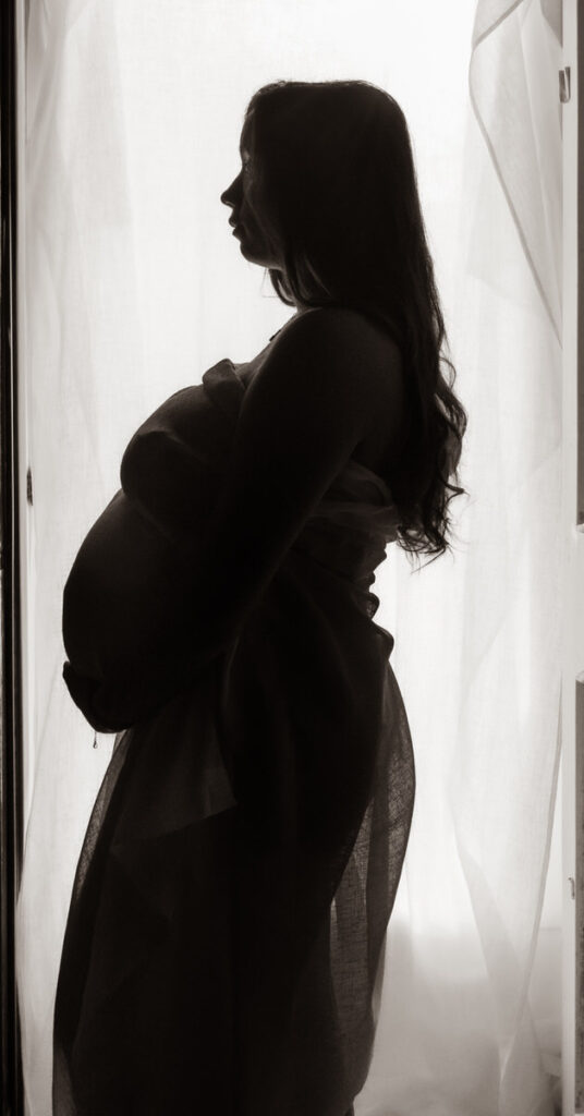 A silhouette of a pregnant woman standing by a sheer curtain, highlighting her profile.