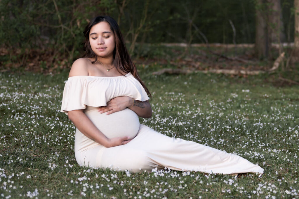 A pregnant woman in an off-shoulder dress sitting in a field of small white flowers, cradling her belly.