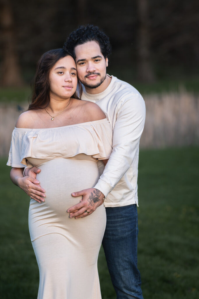 A pregnant woman in an off-shoulder dress being embraced by her partner in a grassy outdoor setting.