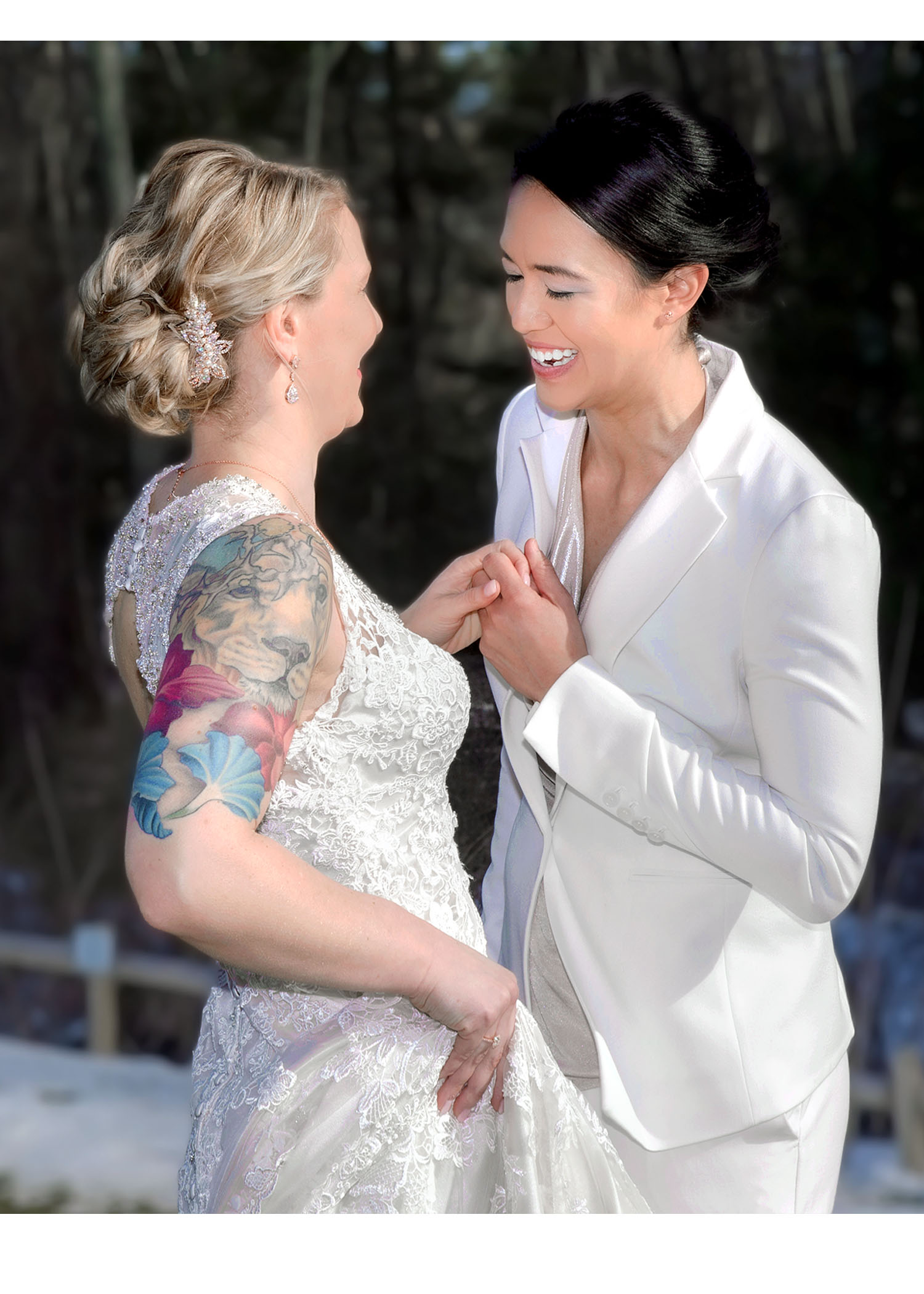 Two brides laughing and holding hands during an outdoor winter wedding, one in a lace dress with a colorful floral tattoo, the other in a white suit. Captured by Boston queer wedding photographer Kathryn Blair Photography.