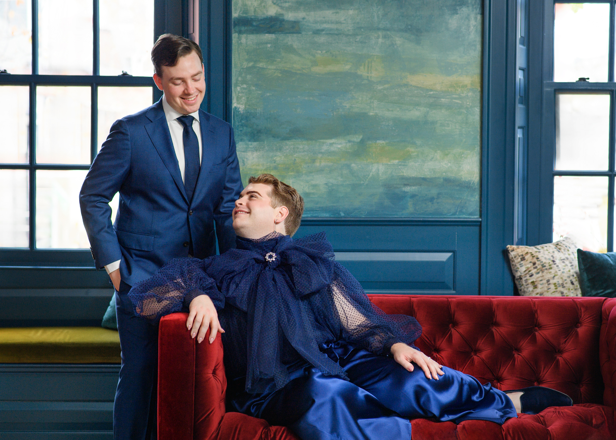 A queer couple during their wedding celebration in Salem, Massachusetts: one in a classic navy suit, the other reclining on a red velvet couch in a flowing blue ensemble. Captured by inclusive wedding photographer Kathryn Blair Photography.